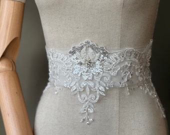 May include: A white lace bridal belt with silver sequins and a pearl embellishment. The belt is designed with a floral pattern and is perfect for adding a touch of elegance to a wedding dress.