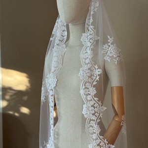 Op de afbeelding: Een witte bruids sluier met een delicate bloemenkant rand. De kant is een gegolfd ontwerp met kleine witte bloemen en kralen.