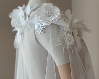 May include: White tulle bridal cape with delicate floral appliques and pearl embellishments. The cape features a large flower on each shoulder.