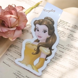 Può includere: Un segnalibro con un'illustrazione di un cartone animato di una donna in un vestito giallo e capelli castani. Il segnalibro ha il testo "Fairyshop". Il segnalibro è su un libro aperto con una rosa rosa sullo sfondo.