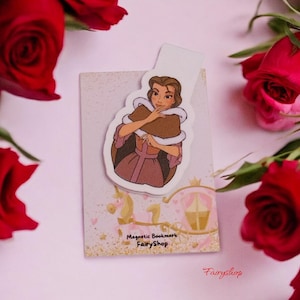 Può includere: Un segnalibro magnetico con un'illustrazione a cartoni animati di una donna con un vestito rosa e uno scialle marrone. Il segnalibro ha il testo "Magnetic Bookmark FairyShop" ed è circondato da rose rosse.