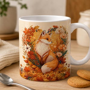 Può includere: Una tazza in ceramica bianca con un'illustrazione centrale di una volpe rossa seduta al centro di una ghirlanda autunnale di foglie d'acero arancioni e marroni, ghiande e vari fiori, con piccoli cuori bianchi su uno sfondo color crema.
