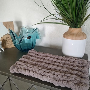 May include: A brown knitted throw pillow with a textured surface sits on a gray dresser with a blue glass vase and a wooden plant stand.