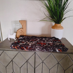 May include: A rectangular, hand-knitted blanket in shades of black, gray, red, and white. The blanket is placed on a gray cabinet with geometric door panels. A white cat figurine and a wooden sign that reads "BE KIND DO GOOD" are also visible.