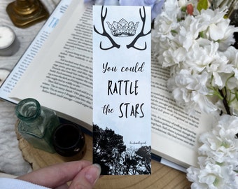 May include: A rectangular paper bookmark featuring a black line art illustration of a crown and antlers at the top. The text reads You could RATTLE the STARS in black script and print. The bottom section displays a black and white forest silhouette.
