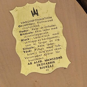 May include: A yellow label with black text detailing the materials used in the construction of a pair of shoes. The label reads "VARUINFORMATION" and lists the materials as "Fullnarvad ungn&ouml;tshud", "Akta skinnfoder", "&Auml;kta l&auml;der", "L&auml;derklack", "H&aring;rdgummi", and "&Auml;kta l&auml;der". The label also includes the company name "AB ALFR. HANSSONS SKOFABRIK BJURS&Aring;S" and the number "40".