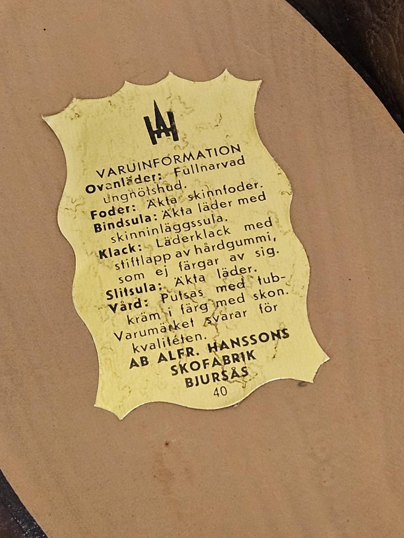 May include: A yellow label with black text detailing the materials used in the construction of a pair of shoes. The label reads "VARUINFORMATION" and lists the materials as "Fullnarvad ungn&ouml;tshud", "Akta skinnfoder", "&Auml;kta l&auml;der", "L&auml;derklack", "H&aring;rdgummi", and "&Auml;kta l&auml;der". The label also includes the company name "AB ALFR. HANSSONS SKOFABRIK BJURS&Aring;S" and the number "40".