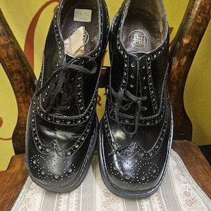 Könnte beinhalten: Ein Paar schwarze Oxford-Schuhe aus Leder mit dekorativen Perforationen und Schnürverschlüssen. Die Schuhe werden auf einer gemusterten Oberfläche präsentiert, die ihr klassisches Design zeigt. Schuhgröße 39.