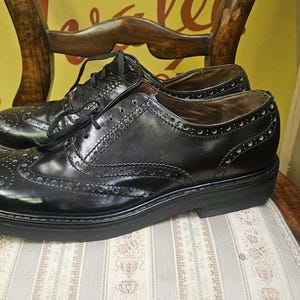 Może przedstawiać: Para czarnych skórzanych butów typu brogue z ozdobnymi perforacjami i grubą podeszwą. Buty są sznurowane i leżą na krześle w wzory. Klasyczny styl.