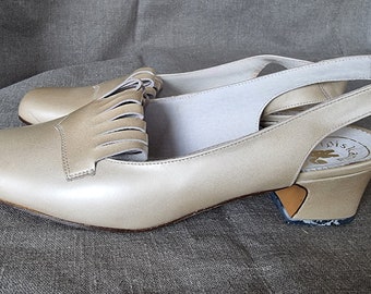 Puede incluir: Un par de zapatos de tacón de aguja beige de cuero con un detalle de flecos en la parte delantera. Los zapatos tienen un tacón bajo de bloque y son de la marca "Tais"
