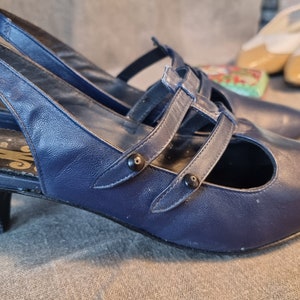 Peut inclure: Une paire d'escarpins bleus marine à bout pointu et petit talon. Les chaussures ont plusieurs brides sur le dessus et une fermeture à boucle. D'autres chaussures sont visibles en arrière-plan.