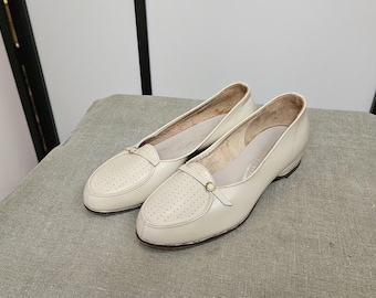 Puede incluir: Un par de mocasines de cuero de color blanco roto con un diseño perforado y un adorno de hebilla dorada.