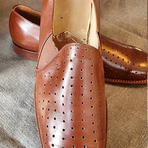 May include: A pair of brown leather slip-on shoes with a perforated design. The shoes have a brown elastic band at the back of the heel.