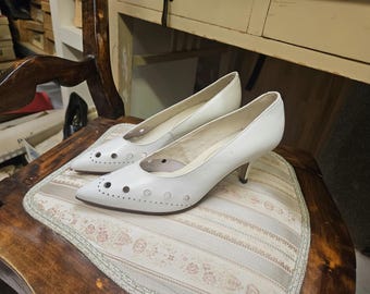 Puede incluir: Un par de zapatos de tacón de cuero blanco con recortes. Los zapatos están sobre una silla de madera marrón claro con un cojín de estampado floral.