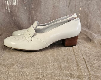 Puede incluir: Zapatos de tacón de cuero blanco con un pequeño detalle de hebilla en la parte delantera. Los zapatos tienen un tacón de madera.