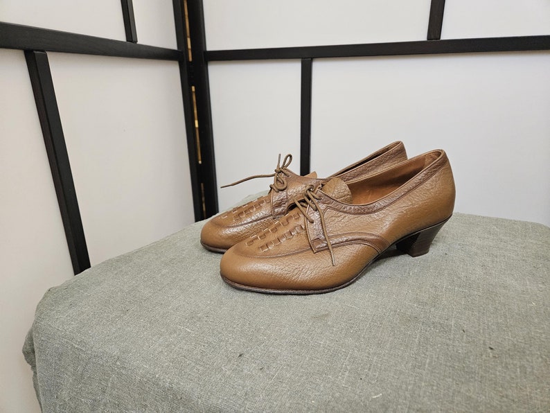 May include: A pair of brown leather shoes with a lace-up design and a low heel. The shoes have a textured leather upper and a smooth leather sole.