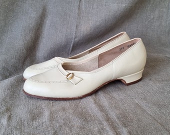May include: A pair of off-white leather shoes with a gold buckle detail. The shoes have a low heel and a pointed toe.