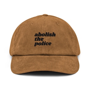 Puede incluir: Una gorra de béisbol de pana marrón con el texto "abolish the police" bordado en negro.