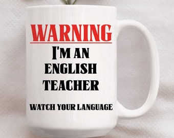 May include: White ceramic mug with a red warning sign that says "WARNING I'M AN ENGLISH TEACHER WATCH YOUR LANGUAGE". The mug is sitting on a white surface with a green leafy branch.