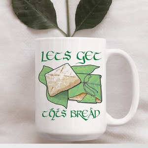 May include: White ceramic mug with a green graphic of two pieces of bread wrapped in green leaves. The text "Let's get this bread" is written in green on the mug.