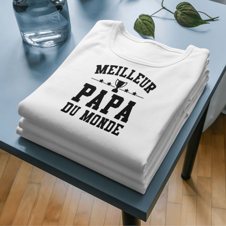 Op de afbeelding: Een wit T-shirt met zwarte tekst die "MEILLEUR PAPA DU MONDE" luidt, met een trofee-icoon boven het woord "PAPA".
