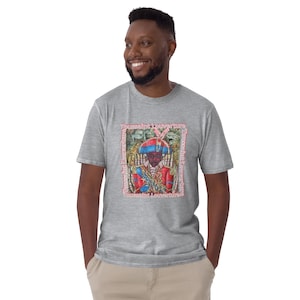 May include: A light gray t-shirt featuring a colorful graphic of a person in a red and blue uniform, with the text "Toussaint Louverture" in a pink frame. The shirt is made of a soft material.