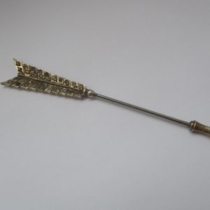 May include: A decorative arrow-shaped pin. It has a silver shaft and a gold-coloured arrowhead with a detailed, patterned design. The pin's end has a small, gold-coloured handle. The pin is set against a plain, light background.
