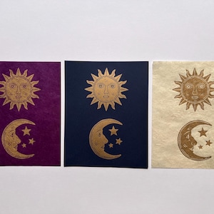 May include: Three celestial-themed art prints. Each print features a gold sun and moon design with stars. The backgrounds are deep purple, navy blue, and cream-colored, respectively. The sun and moon are detailed with facial features.