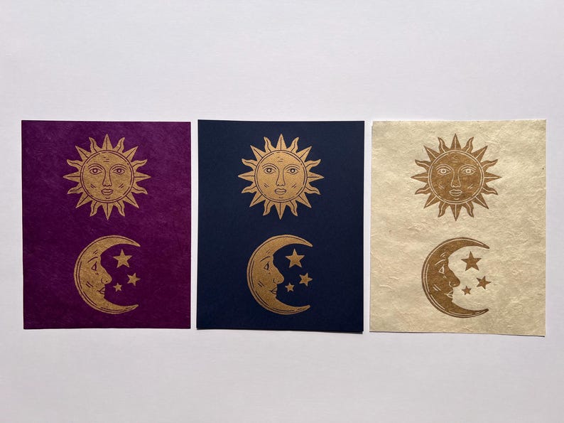 May include: Three celestial-themed art prints. Each print features a gold sun and moon design with stars. The backgrounds are deep purple, navy blue, and cream-colored, respectively. The sun and moon are detailed with facial features.