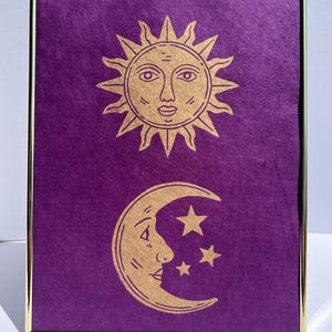 May include: Framed artwork featuring a gold sun and moon design on a textured purple background. The sun has a face, and the moon is a crescent with stars. The frame is gold.