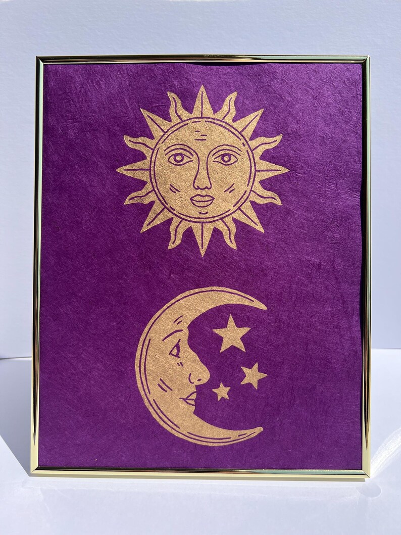 May include: Framed artwork featuring a gold sun and moon design on a textured purple background. The sun has a face, and the moon is a crescent with stars. The frame is gold.