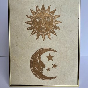 May include: A framed print featuring a gold sun and moon design. The sun has a face and rays, while the moon has a face and three stars. The artwork is on textured paper and set in a gold frame.