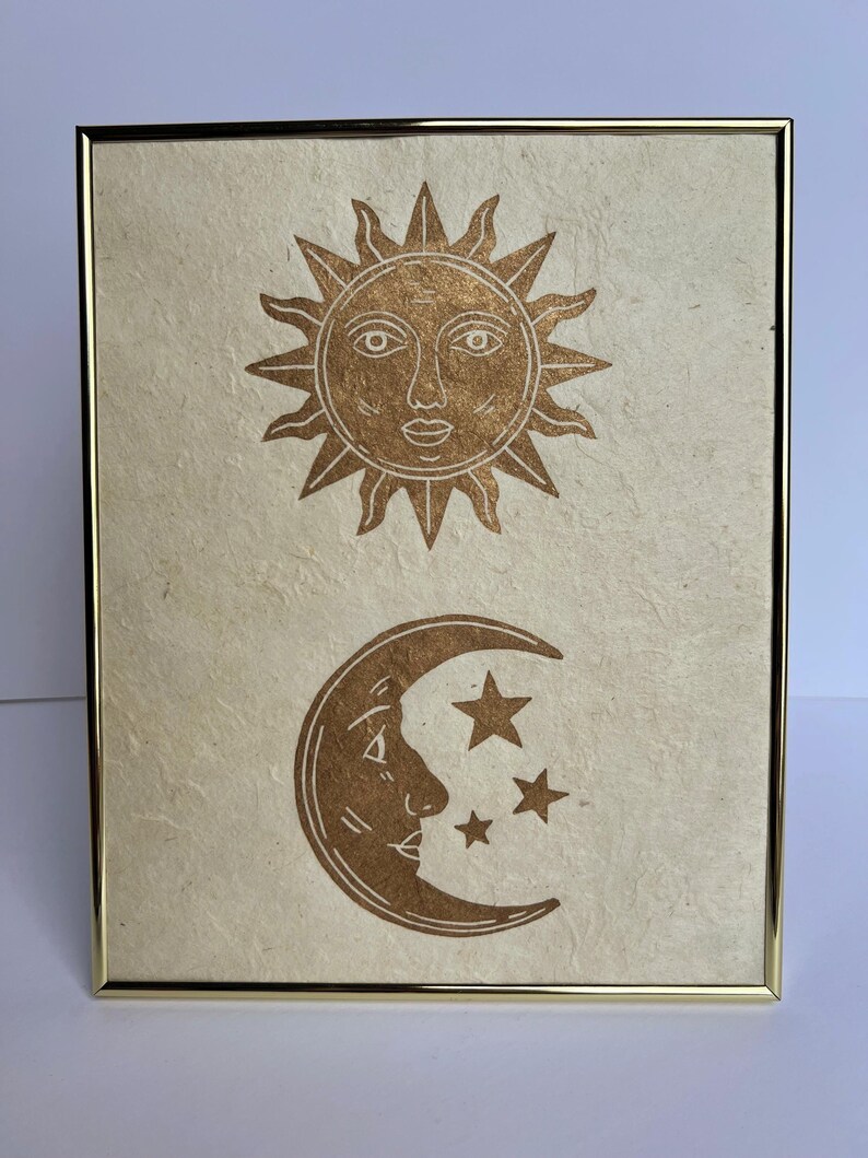 May include: A framed print featuring a gold sun and moon design. The sun has a face and rays, while the moon has a face and three stars. The artwork is on textured paper and set in a gold frame.