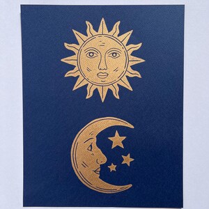 May include: A navy blue card features gold illustrations of celestial bodies. The top shows a sun with a face and spiky rays. Below, a crescent moon with a face and three stars are depicted. The design is simple and elegant.