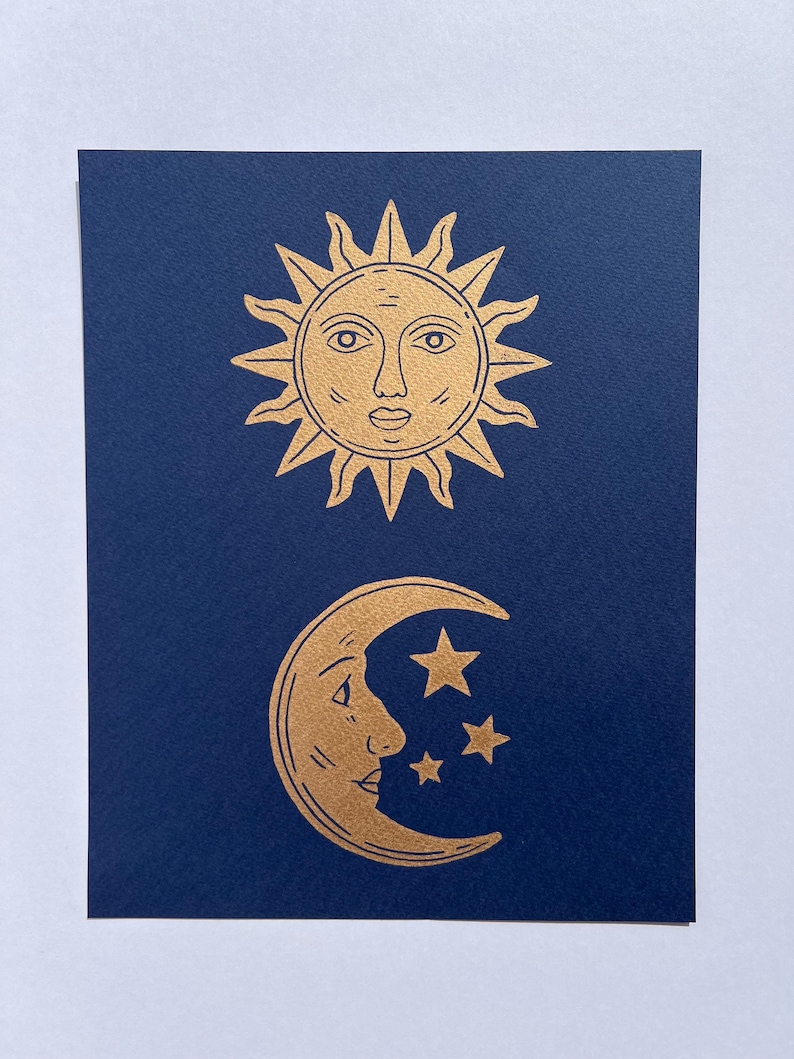 May include: A navy blue card features gold illustrations of celestial bodies. The top shows a sun with a face and spiky rays. Below, a crescent moon with a face and three stars are depicted. The design is simple and elegant.