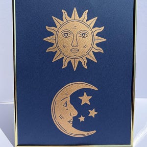 May include: Framed art print featuring a gold sun and moon design on a navy blue background. The sun has a face and the moon is a crescent with three stars. The frame is gold.