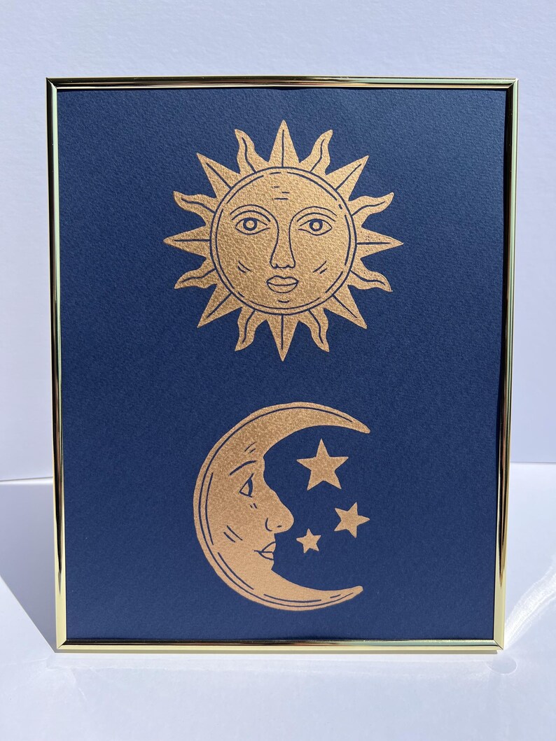 May include: Framed art print featuring a gold sun and moon design on a navy blue background. The sun has a face and the moon is a crescent with three stars. The frame is gold.