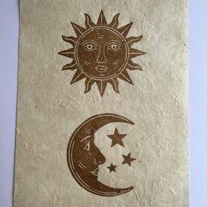 May include: A tan-colored paper print featuring a sun and moon design. The sun is at the top with a face and rays, while the moon with a face and stars is below. The print has a textured, handmade appearance.
