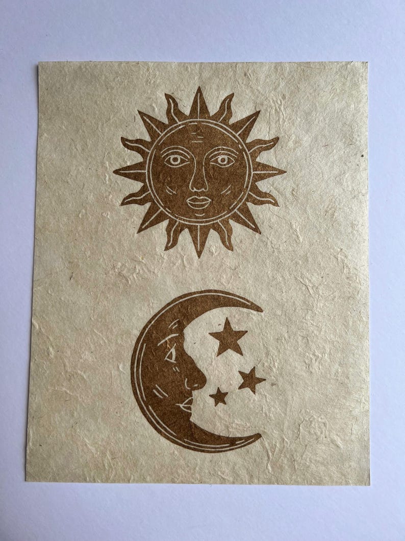 May include: A tan-colored paper print featuring a sun and moon design. The sun is at the top with a face and rays, while the moon with a face and stars is below. The print has a textured, handmade appearance.