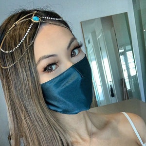 May include: A woman with long brown hair wearing a teal blue satin face mask and a gold chain headpiece with a turquoise stone. She is wearing a white lace top and a silver necklace with a pearl pendant.