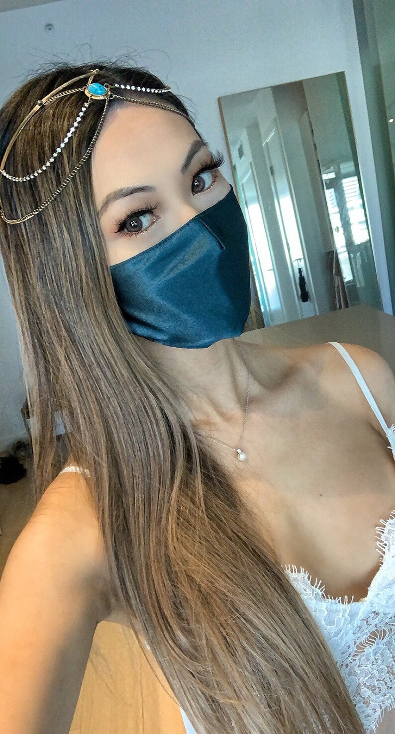 May include: A woman with long brown hair wearing a teal blue satin face mask and a gold chain headpiece with a turquoise stone. She is wearing a white lace top and a silver necklace with a pearl pendant.