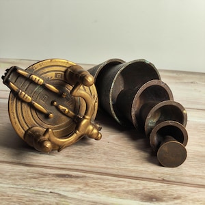 May include: A collection of antique brass measuring tools, including a circular scale with a handle and a set of nested conical measuring cups. The scale has the numbers "23" and "102" on it. The items are on a wooden surface.