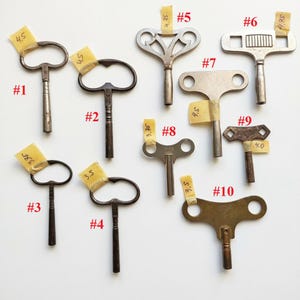 以下が含まれることがあります： 1番から10番まで番号が振られた10本の金属製時計用巻き鍵のコレクション。それぞれ独特な形状の持ち手と軸を持っています。いくつかの鍵には、手書きでcm単位の寸法が記された小さな黄色い粘着テープが貼られています。