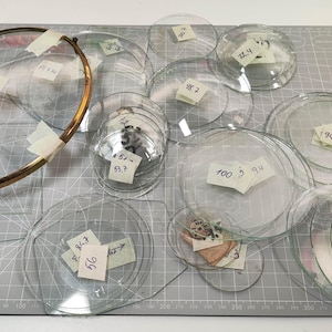 May include: A collection of clear glass lenses and circular objects, some with attached paper labels. A gold-colored metal ring is taped to a clear glass lens. The items are arranged on a gray cutting mat.
