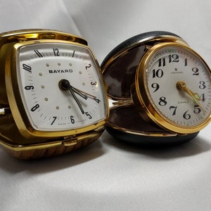 Può includere: Due sveglie da viaggio vintage. Una è quadrata con cornice dorata e quadrante bianco, l'altra è rotonda con cornice dorata e quadrante bianco. Entrambi hanno lancette nere e numeri arabi. L'orologio quadrato indica "BAYARD" e l'orologio rotondo "Lecoultre".