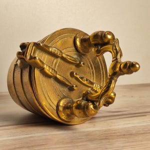 May include: A gold-coloured, antique-style decorative object. The piece features a circular design with a handle and several decorative elements. The object appears to be made of metal and has a vintage aesthetic.