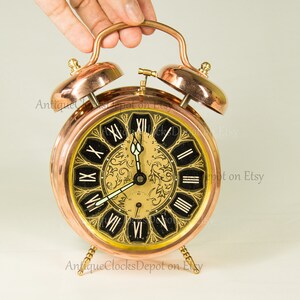 Könnte beinhalten: Ein antiker Kupfer-Wecker mit römischen Ziffern auf einem schwarzen Zifferblatt. Die Uhr hat einen goldfarbenen Hintergrund mit einem floralen Design und zwei Kupferglocken oben.