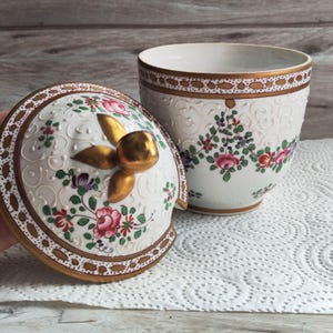 May include: A white porcelain container with a lid, featuring floral designs in pink, purple, and green. The lid has a gold-colored handle and trim. The container has a matching floral pattern and gold trim.