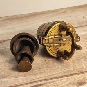 May include: Two antique brass objects are displayed on a wooden surface. One is a cylindrical object with a handle and a circular base. The other is a smaller, cone-shaped object with a circular base. Both objects have a tarnished, aged appearance.