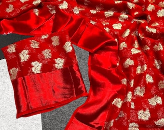 May include: A vibrant red fabric with a gold floral pattern. The material is draped, showcasing its flowing texture. The image includes a matching red sleeve, suggesting a garment like a saree or dress.